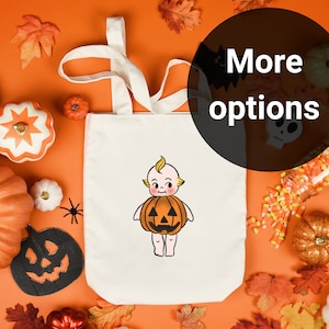 May include: A cream-colored tote bag featuring a cartoon baby dressed as a jack-o'-lantern. The bag has long handles and is surrounded by Halloween decorations, including pumpkins, leaves, and candy corn. The text "More options" is in a black circle.
