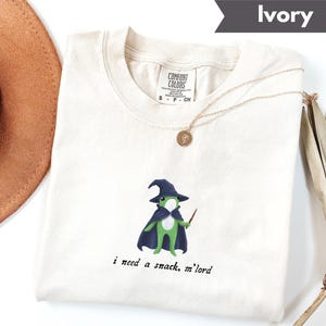May include: Ivory-colored t-shirt with a crew neck. The shirt features a cartoon frog wearing a wizard hat and cape, holding a wand. The text "i need a snack, m'lord" is printed below the frog. A gold necklace is draped across the shirt.