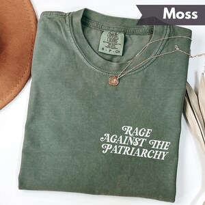 May include: A moss green t-shirt with the text "Rage Against The Patriarchy" in white script. A gold-colored necklace with a pendant rests on the shirt. The shirt is folded and displayed with a brown hat and dried leaves.