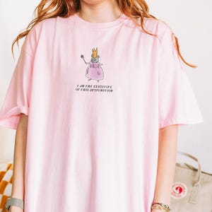 K&ouml;nnte beinhalten: Hellrosa T-Shirt mit einer Cartoon-Maus, die eine Krone und ein Kleid tr&auml;gt und einen Zauberstab h&auml;lt. Der Text "I AM THE EXECUTIVE OF THIS DYSFUNCTION" ist darunter gedruckt. Das T-Shirt hat kurze &Auml;rmel.