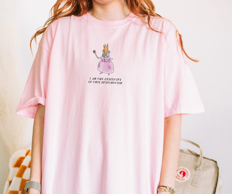 K&ouml;nnte beinhalten: Hellrosa T-Shirt mit einer Cartoon-Maus, die eine Krone und ein Kleid tr&auml;gt und einen Zauberstab h&auml;lt. Der Text "I AM THE EXECUTIVE OF THIS DYSFUNCTION" ist darunter gedruckt. Das T-Shirt hat kurze &Auml;rmel.
