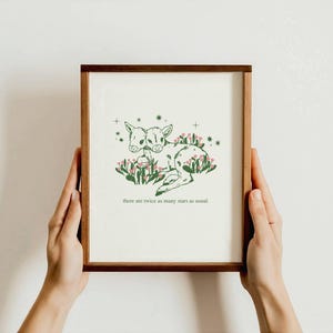 Könnte beinhalten: Ein gerahmter Kunstdruck mit einer Illustration eines zweiköpfigen Hirsches in Grün und Weiß, umgeben von Blumen und Sternen. Der Text "there are twice as many stars as usual" steht darunter. Der Rahmen ist dunkelbraun.