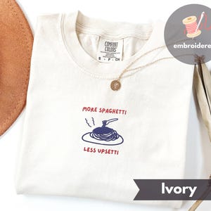 May include: Ivory t-shirt with the embroidered text "MORE SPAGHETTI LESS UPSETTI" in red and blue. The shirt features a spaghetti graphic and a Comfort Colors tag. A gold necklace is visible.