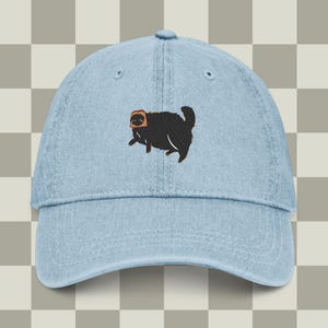 May include: Light blue denim baseball cap featuring an embroidered black cat wearing a piece of toast on its head. The cap has a curved brim and an adjustable strap at the back. The cat design is a whimsical and playful element.