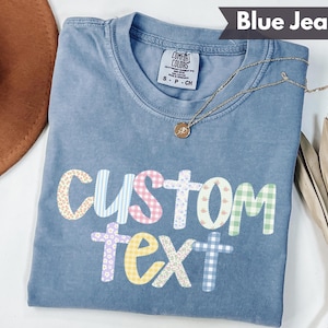 May include: A blue jean Comfort Colors t-shirt with the words "CUSTOM TEXT" in a patchwork design. The patchwork includes floral, gingham, and striped patterns in pastel colors. A gold necklace with a pendant is draped across the shirt.