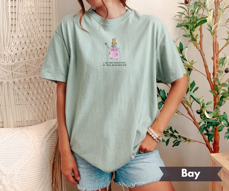 K&ouml;nnte beinhalten: Hellgr&uuml;nes T-Shirt mit einer Cartoon-Maus in einem rosa Kleid und Krone, die einen Zauberstab h&auml;lt. Der Text "I AM THE EXECUTIVE OF THIS DYSFUNCTION" ist darunter aufgedruckt. Das Shirt wird mit Jeansshorts getragen.