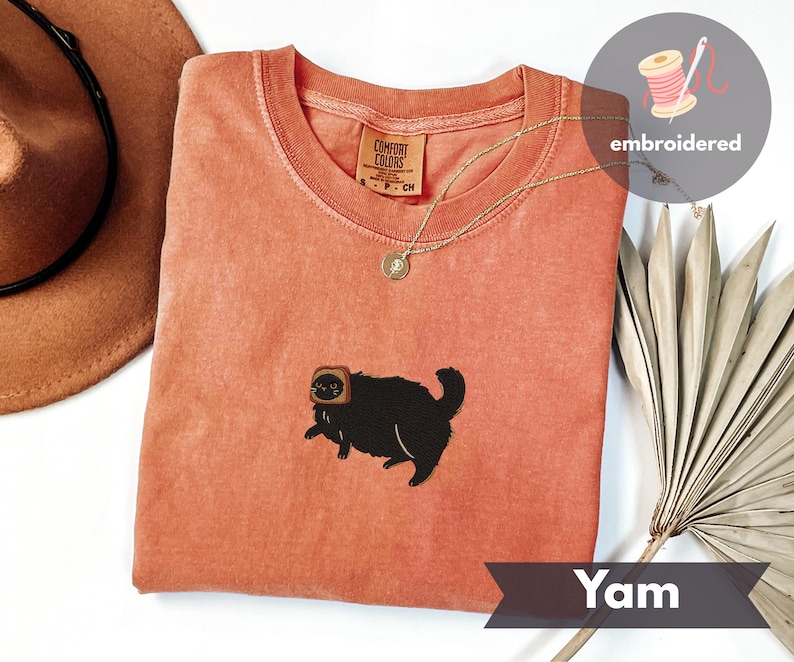 Puede incluir: Una camiseta de color coral con cuello redondo y un gato negro bordado que lleva una rebanada de pan tostado en la cabeza. La camiseta tiene una peque&ntilde;a etiqueta que dice "Comfort Colors". Un collar de oro est&aacute; drapeado sobre la camiseta. La palabra "Yam" est&aacute; impresa en una pancarta.