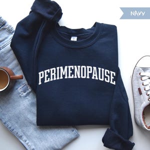 May include: Navy blue sweatshirt with the word "PERIMENOPAUSE" in white block letters. The sweatshirt is paired with a pair of blue jeans, a coffee mug, and a gray sneaker.