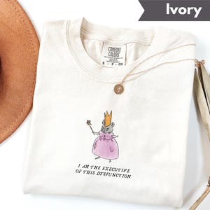 K&ouml;nnte beinhalten: Elfenbeinfarbenes T-Shirt mit einer Illustration einer Maus mit Krone und rosa Kleid, die einen Zauberstab h&auml;lt. Der Text "I AM THE EXECUTIVE OF THIS DYSFUNCTION" ist darunter gedruckt. Eine goldene Halskette liegt auf dem T-Shirt.