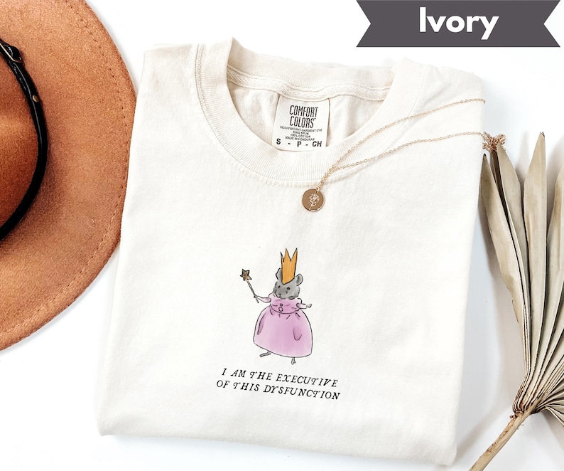 K&ouml;nnte beinhalten: Elfenbeinfarbenes T-Shirt mit einer Illustration einer Maus mit Krone und rosa Kleid, die einen Zauberstab h&auml;lt. Der Text "I AM THE EXECUTIVE OF THIS DYSFUNCTION" ist darunter gedruckt. Eine goldene Halskette liegt auf dem T-Shirt.