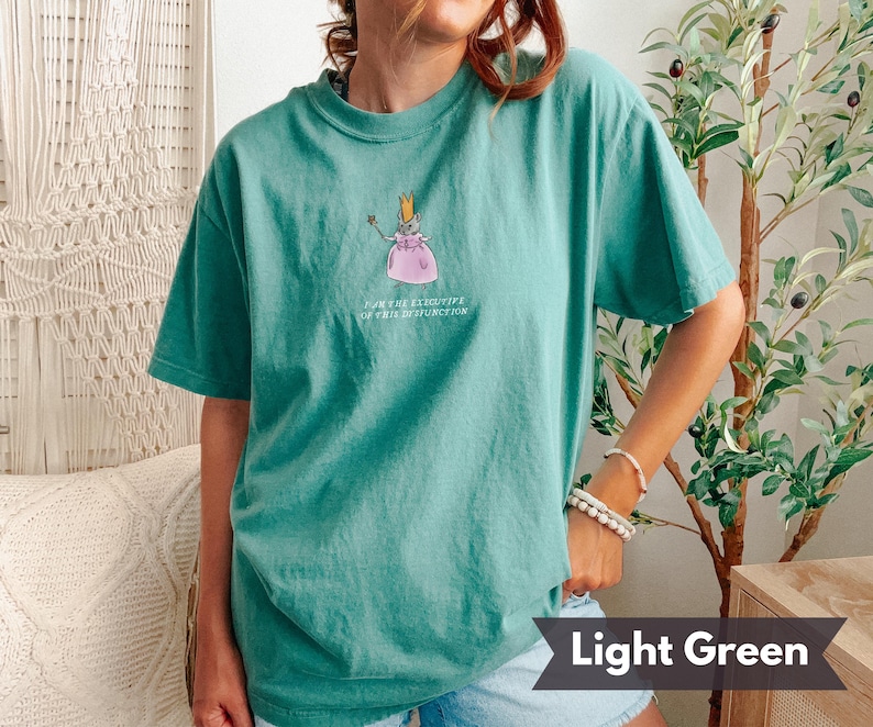 K&ouml;nnte beinhalten: Hellgr&uuml;nes T-Shirt mit einer Cartoon-Illustration einer Maus mit Krone und rosa Kleid, die einen Zauberstab h&auml;lt. Der Text auf dem Shirt lautet "I AM THE EXECUTIVE OF THIS DYSFUNCTION".