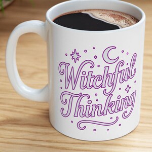 May include: White ceramic mug with the words "Witchful Thinking" in purple script, along with a crescent moon, stars, and dots. The mug is filled with dark coffee and sits on a wooden surface.