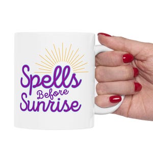May include: White ceramic mug with the text "Spells Before Sunrise" in purple script, with a gold sunburst design. The mug is held by a hand with red nail polish.