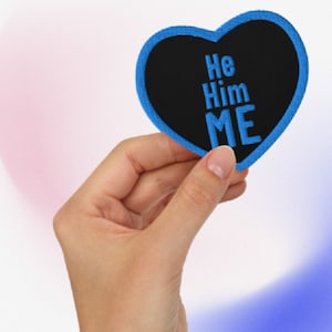 May include: A blue heart-shaped patch with black fabric and blue embroidery that reads "He Him Me."