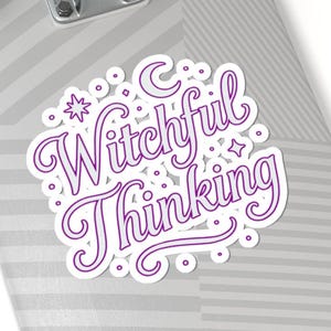 May include: A white sticker with the text "Witchful Thinking" in purple script. The sticker has a white border and is decorated with stars, dots, and a crescent moon. The sticker is on a skateboard.