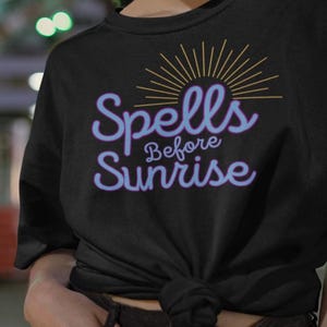 May include: Black t-shirt featuring the text "Spells Before Sunrise" in a light blue script with a sunburst design above. The shirt is knotted at the waist.