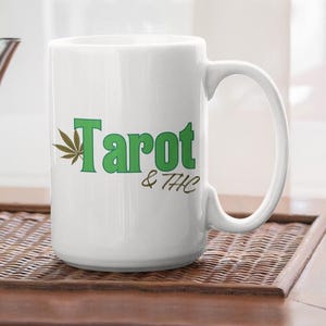Op de afbeelding: Witte keramische mok met de woorden "Tarot & THC" in het groen, met een cannabisblad-afbeelding. Een zilveren theepot staat naast de mok op een houten dienblad.
