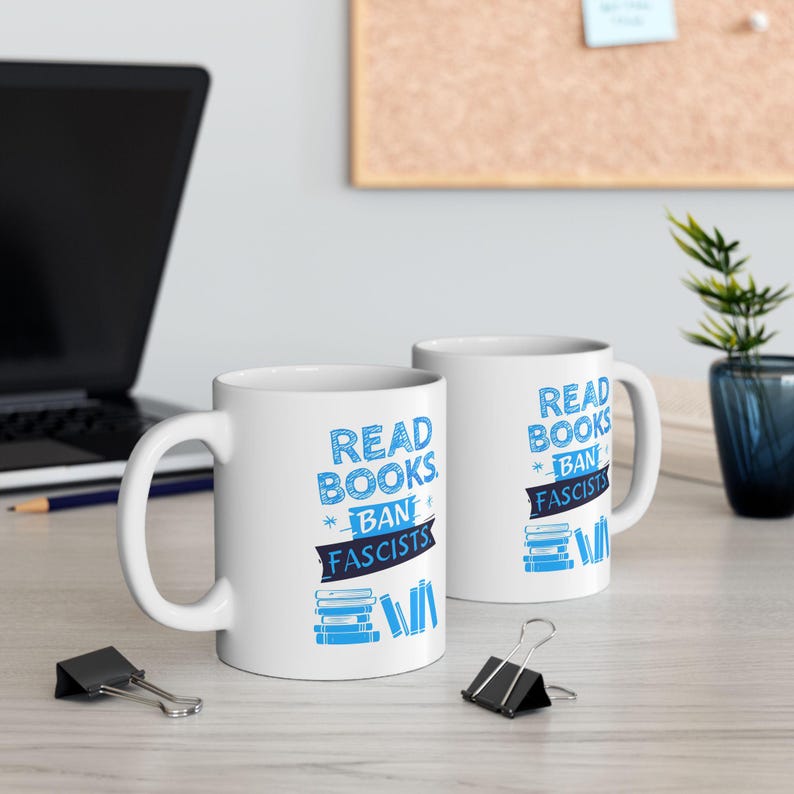 ceramic mug featuring bold blue hand-drawn text that reads "Read Books Ban Fascists" with illustrated books beneath the text.