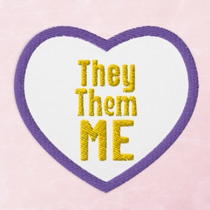 May include: A white heart-shaped patch with a purple outline. The text "They Them ME" is embroidered in yellow.