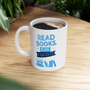 ceramic mug featuring bold blue hand-drawn text that reads "Read Books Ban Fascists" with illustrated books beneath the text.