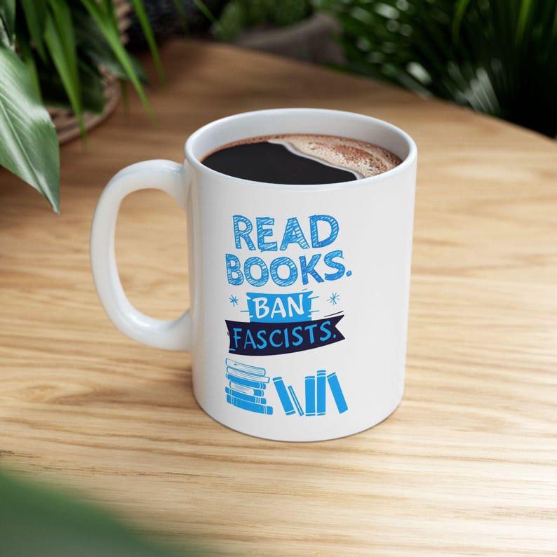 ceramic mug featuring bold blue hand-drawn text that reads "Read Books Ban Fascists" with illustrated books beneath the text.