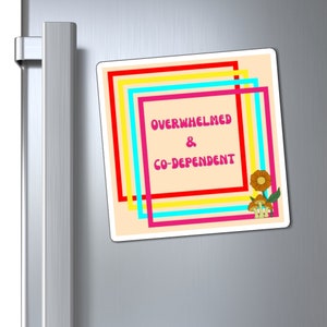 May include: A square magnet with the words "OVERWHELMED & CO-DEPENDENT" in pink text, framed by colorful lines. A flower and mushroom graphic is in the bottom right corner. The magnet is attached to a silver refrigerator door.