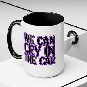 May include: White ceramic mug with a black interior and handle. The mug features the text "WE CAN CRY IN THE CAR" in a purple and black design. The mug is on a white surface.