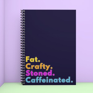 Op de afbeelding: Een donkerblauw spiraalnotitieblok met de tekst "Fat. Crafty. Stoned. Caffeinated." in kleurrijke letters.