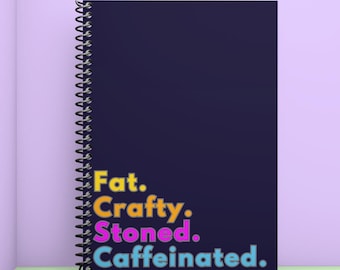Pode incluir: Um caderno de espiral azul escuro com o texto "Fat. Crafty. Stoned. Caffeinated." em letras coloridas.
