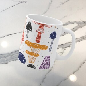 White ceramic mug featuring colorful illustrations of various mushrooms, perfect for mushroom enthusiasts and those who love nature-inspired designs.