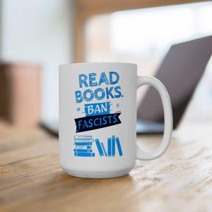 ceramic mug featuring bold blue hand-drawn text that reads "Read Books Ban Fascists" with illustrated books beneath the text.