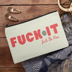 May include: Cream canvas zipped pouch featuring the words "FUCK IT" in coral block letters, a blue daisy, and the phrase "Just Be Nice." The pouch is on a wooden surface with gold hair clips.