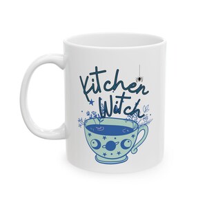 Puede incluir: Taza de cerámica blanca con una ilustración azul y verde de una taza de té con estrellas y lunas. El texto "Kitchen Witch" está escrito en cursiva azul sobre la taza de té.