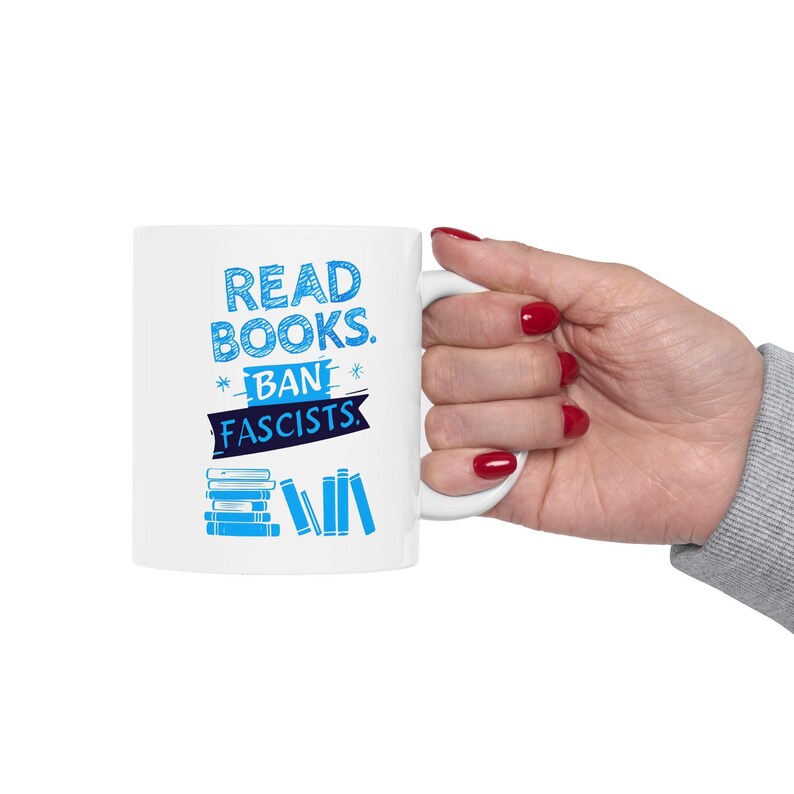 ceramic mug featuring bold blue hand-drawn text that reads "Read Books Ban Fascists" with illustrated books beneath the text.