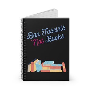 May include: A spiral-bound notebook with a black cover. The cover features the text "Ban Fascists Not Books" in a neon-style font. Below the text is a stack of illustrated books in various colors.