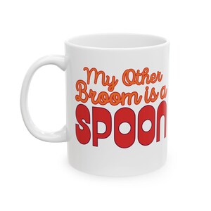 Puede incluir: Taza de cerámica blanca con el texto "My Other Broom is a Spoon" en letras naranjas y rojas. La taza tiene un asa curva y un acabado brillante. El diseño está centrado en la taza.