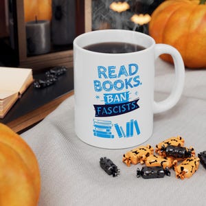 ceramic mug featuring bold blue hand-drawn text that reads "Read Books Ban Fascists" with illustrated books beneath the text.