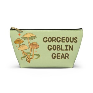 T-bottom pouch in moss green with a mushroom and the words Gorgeous Goblin Gear on the front and a cute snail with a positive message of You're doing great! on the back. White zipper