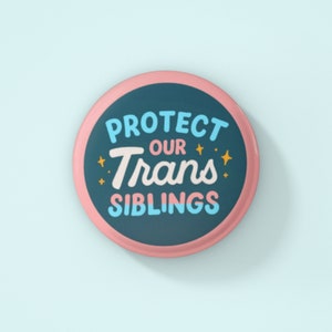 May include: A pink and blue button with the text "Protect Our Trans Siblings" in white lettering with gold stars.