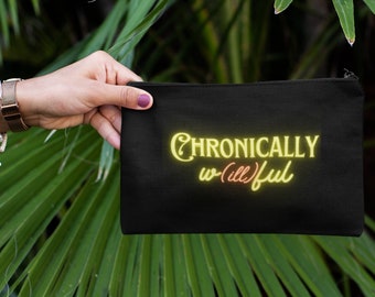 Pode incluir: Bolsa preta com zíper com o texto "Chronically Willful" em uma fonte em estilo neon. O texto é amarelo e rosa. A bolsa é segurada contra um fundo de folhas verdes.
