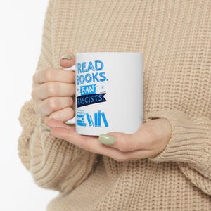 ceramic mug featuring bold blue hand-drawn text that reads "Read Books Ban Fascists" with illustrated books beneath the text.