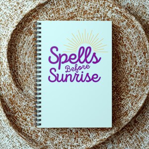 May include: A spiral-bound notebook with a light blue cover. The cover features the words "Spells Before Sunrise" in purple script with a gold sunburst design above. The notebook rests on a textured, woven surface.