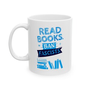 ceramic mug featuring bold blue hand-drawn text that reads "Read Books Ban Fascists" with illustrated books beneath the text.
