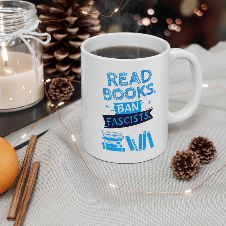 ceramic mug featuring bold blue hand-drawn text that reads "Read Books Ban Fascists" with illustrated books beneath the text.