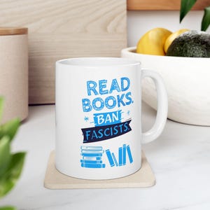 ceramic mug featuring bold blue hand-drawn text that reads "Read Books Ban Fascists" with illustrated books beneath the text.