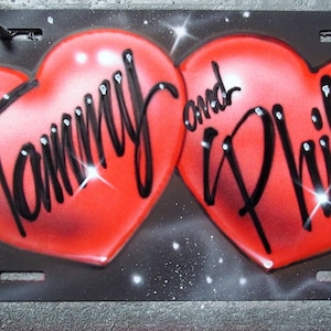 May include: Two red hearts with the names "Tammy and Phil" written in black cursive on a black background with white glitter.