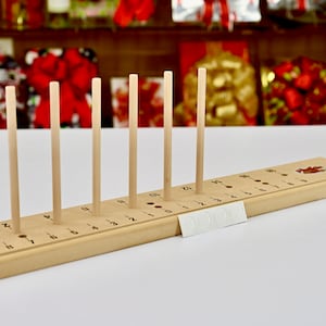 May include: A wooden bow making tool with eight dowels and a numbered scale. The tool is designed to help crafters create bows of different sizes.