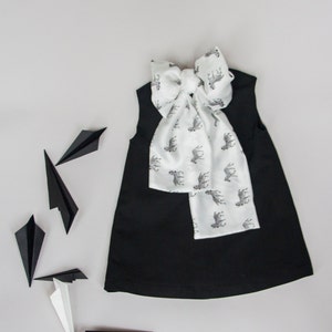 May include: A black sleeveless dress with a white zebra print bow tie. The dress is laying flat on a white surface with several black paper airplanes scattered around it.