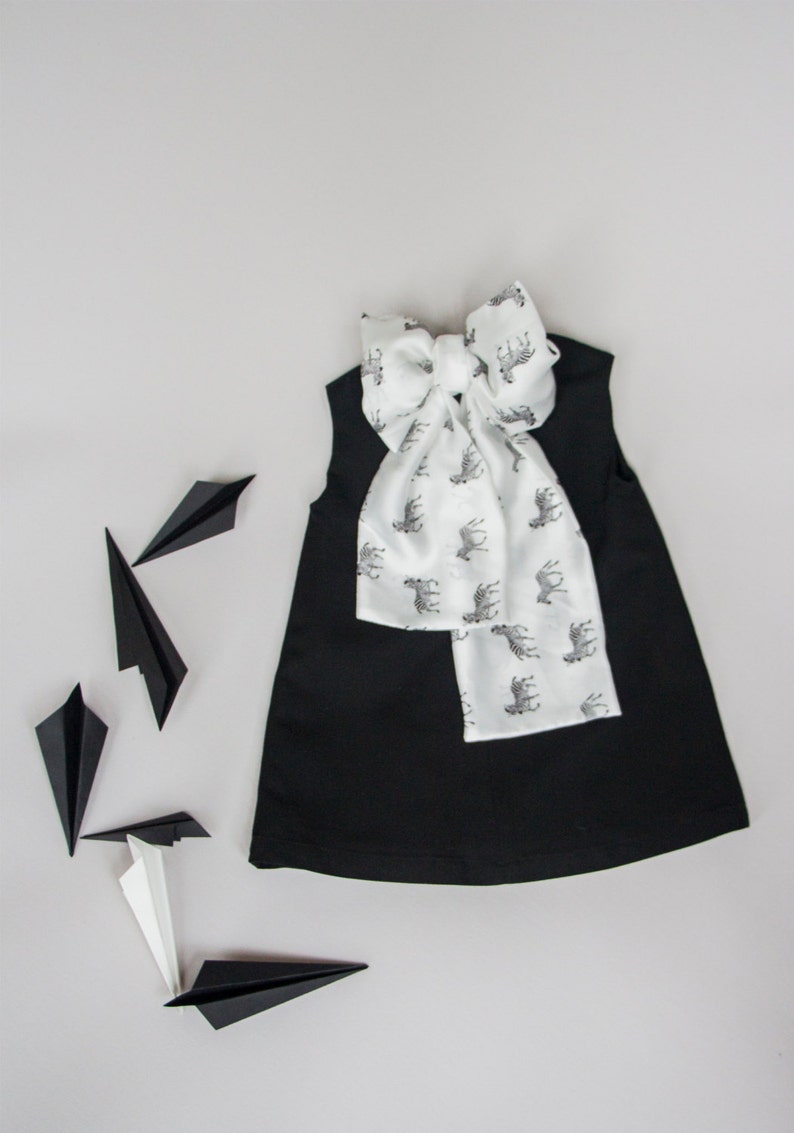 May include: A black sleeveless dress with a white zebra print bow tie. The dress is laying flat on a white surface with several black paper airplanes scattered around it.