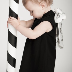 May include: A young child wearing a black sleeveless dress with a white bow tied at the back. The bow has a zebra print pattern. The child is holding onto a black and white striped pole.